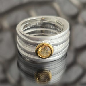 Puede incluir: Un anillo de plata con un bisel dorado que engarza una gema redonda de color amarillo claro. El anillo tiene un diseño multibanda con un acabado pulido, que refleja la luz. La gema es el punto focal.