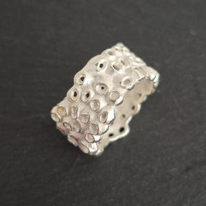 Könnte beinhalten: Ein silberner Ring mit einer strukturierten Oberfläche, die mit zahlreichen kleinen, unregelmäßigen Löchern versehen ist. Der Ring hat ein breites Band und ein mattes Finish, was ihm ein einzigartiges, organisches Aussehen verleiht. Der Ring wird auf dunklem Hintergrund präsentiert.