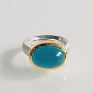 Könnte beinhalten: Ein silberner Ring mit einem goldenen Band und einem türkisfarbenen ovalen Edelstein.