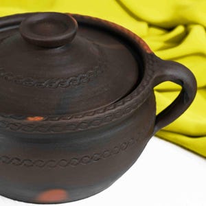 May include: A brown clay pot with a lid and two handles. The pot has a decorative design around the rim.