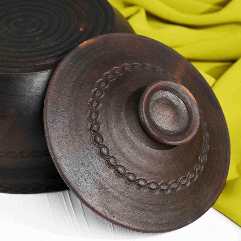 May include: A brown ceramic pot with a lid. The lid has a decorative design around the edge and a knob in the center.
