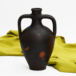 Puede incluir: Un jarrón de cerámica marrón oscuro con dos asas y un cuello estrecho. El jarrón tiene una superficie texturizada con acentos rojo-anaranjados. El jarrón está sobre un fondo blanco con una tela verde.