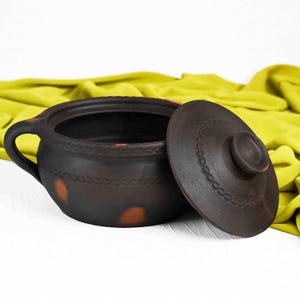 May include: A brown clay pot with a lid. The pot has a decorative design around the rim and a few brown spots on the side. The lid has a knob on top.