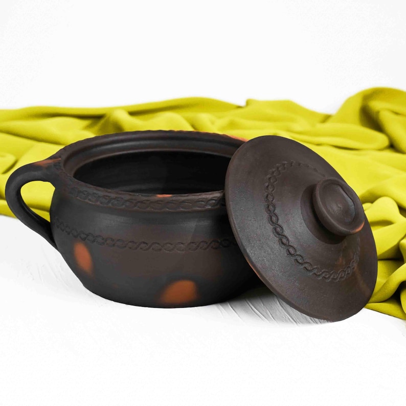 May include: A brown clay pot with a lid. The pot has a decorative design around the rim and a few brown spots on the side. The lid has a knob on top.