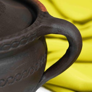 May include: A dark brown clay pot with a lid and two handles. The pot has a decorative design around the rim and body.
