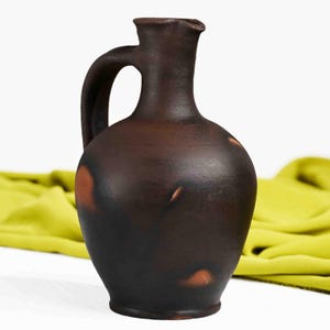 May include: A dark brown ceramic jug with a handle and a spout. The jug has a rustic appearance with reddish-brown spots and a smooth surface. A yellow fabric is in the background.