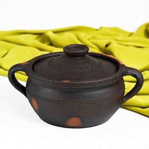 May include: A brown ceramic pot with a lid and two handles. The pot has a decorative design around the rim and a few brown spots on the side.