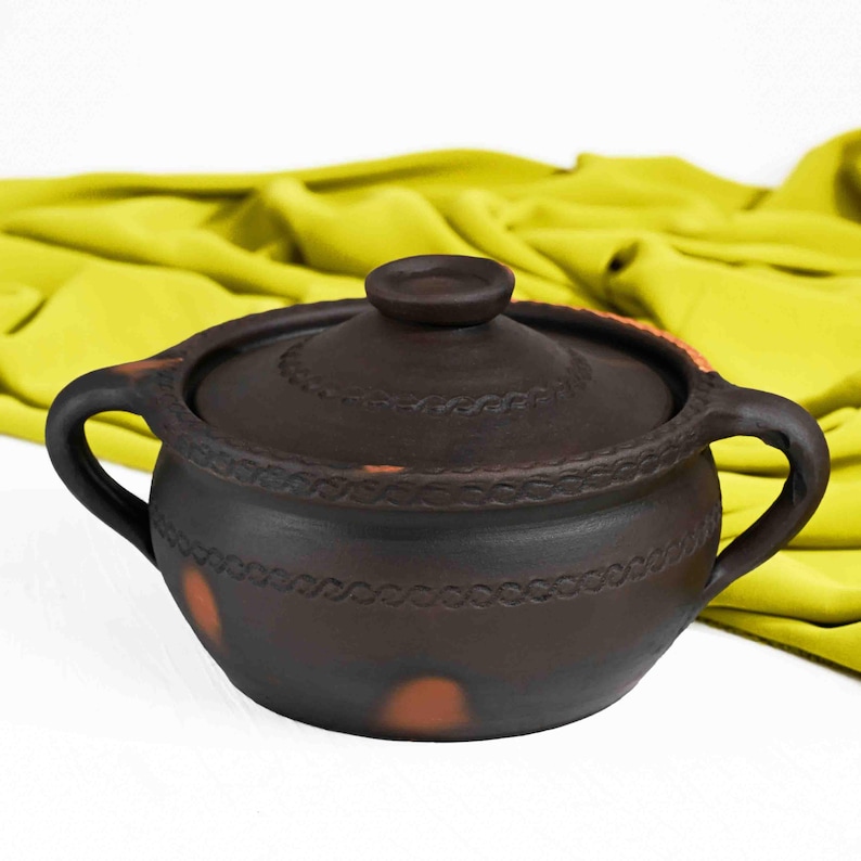 May include: A brown ceramic pot with a lid and two handles. The pot has a decorative design around the rim and a few brown spots on the side.