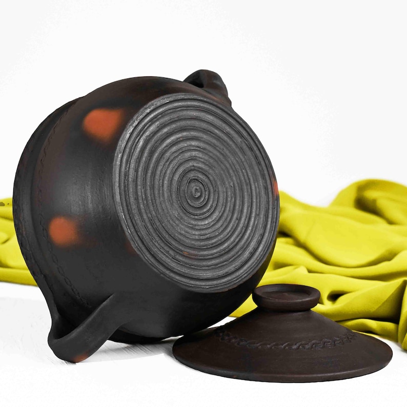 May include: A black clay pot with a lid. The pot has a textured, circular design on the bottom and orange spots on the side. The lid has a braided design around the edge.