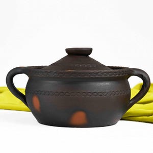 May include: A brown clay pot with a lid and two handles. The pot has a decorative design around the rim and a few brown spots on the side.