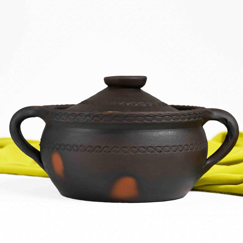 May include: A brown clay pot with a lid and two handles. The pot has a decorative design around the rim and a few brown spots on the side.