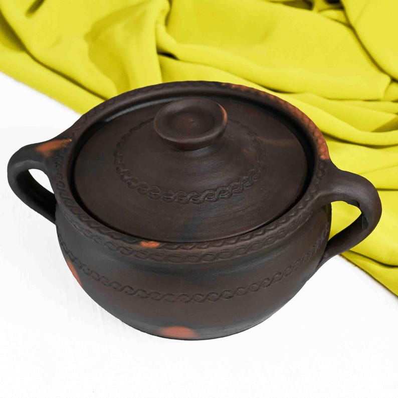 May include: A brown clay pot with a lid and two handles. The pot has a decorative design around the rim.