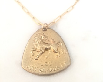 May include: A gold-toned necklace featuring a triangular pendant with a raised lion figure, the Leo zodiac symbol, and the text LEO JULY 24 - AUG 22. The pendant hangs from a delicate gold-toned chain with elongated rectangular links.