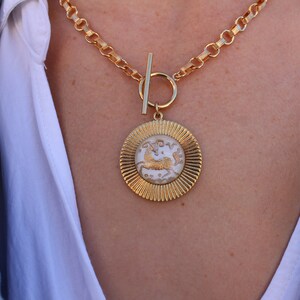May include: A gold chain necklace with a large, round pendant featuring a white, detailed design of a mythical creature. The pendant has a textured, sunburst-like border.