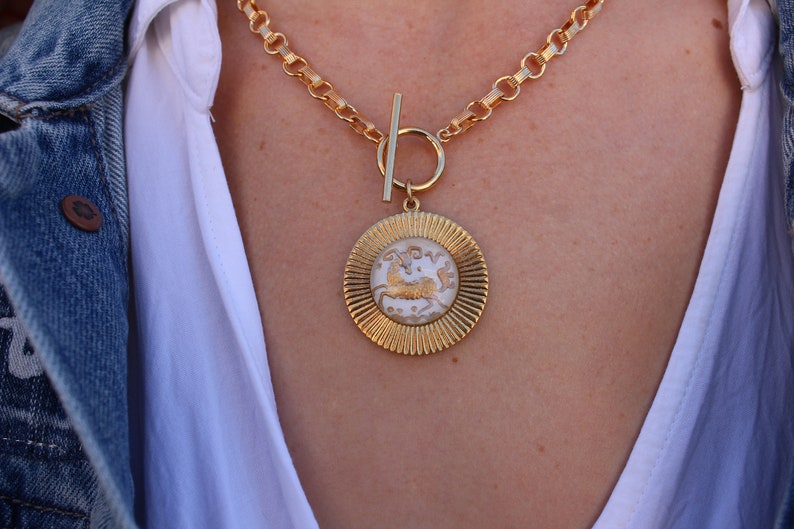 May include: A gold chain necklace with a large, round pendant featuring a white, detailed design of a mythical creature. The pendant has a textured, sunburst-like border.