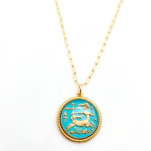 May include: A gold-toned necklace featuring a round pendant with a bright turquoise enamel background. The pendant displays a raised gold-coloured centaur figure holding a bow and arrow, surrounded by a textured, beaded gold-toned border.