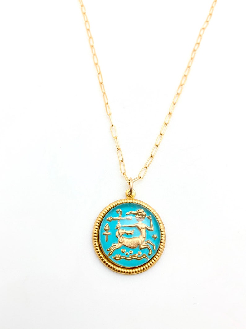 May include: A gold-toned necklace featuring a round pendant with a bright turquoise enamel background. The pendant displays a raised gold-coloured centaur figure holding a bow and arrow, surrounded by a textured, beaded gold-toned border.