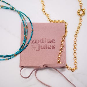 May include: A dusty pink square pouch with the text zodiac jules printed in dark pink, featuring a turquoise beaded necklace with gold accents and a gold chain necklace with a circular clasp resting on top.