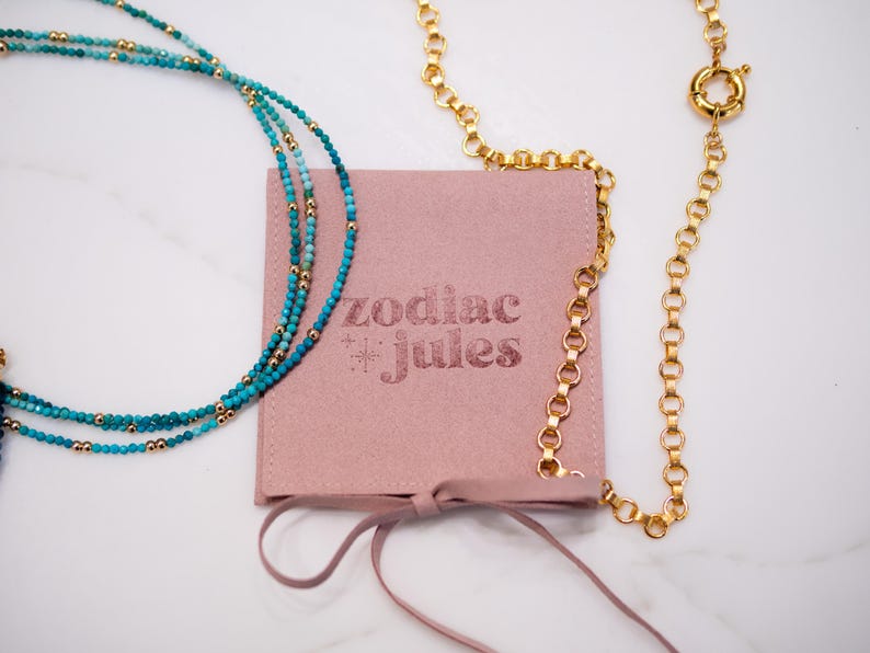 May include: A dusty pink square pouch with the text zodiac jules printed in dark pink, featuring a turquoise beaded necklace with gold accents and a gold chain necklace with a circular clasp resting on top.