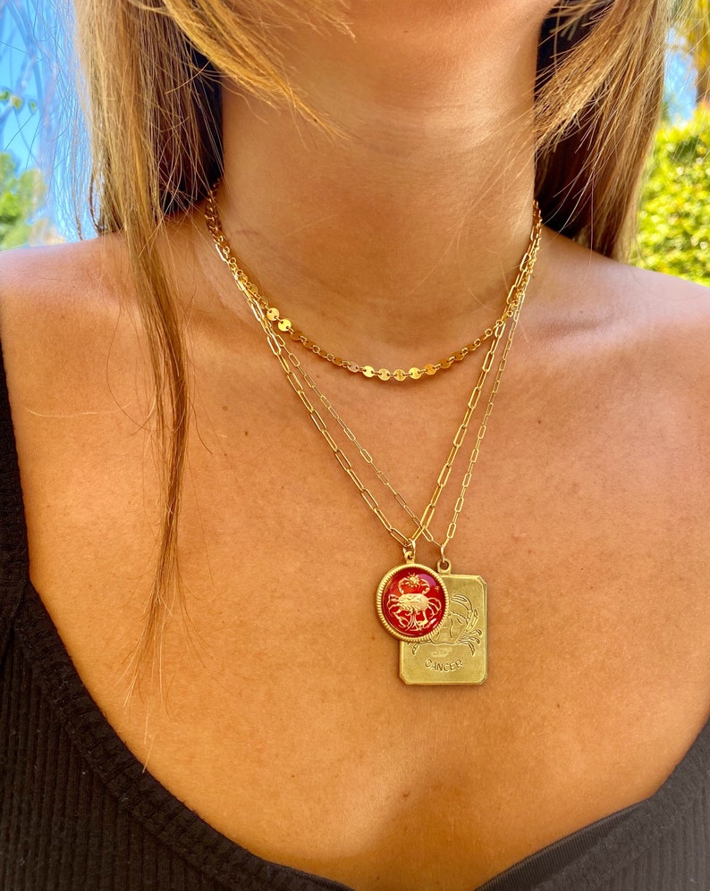 May include: A gold chain necklace with a red and gold zodiac pendant featuring the Cancer symbol. The pendant is a round charm with a red background and a gold crab design. The pendant is attached to a rectangular tag with the word "Cancer" engraved on it.
