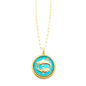 May include: A gold-tone necklace featuring a round pendant with a bright turquoise enamel inlay. The pendant displays two embossed gold fish swimming in opposite directions, representing the Pisces zodiac sign, with a textured gold border.