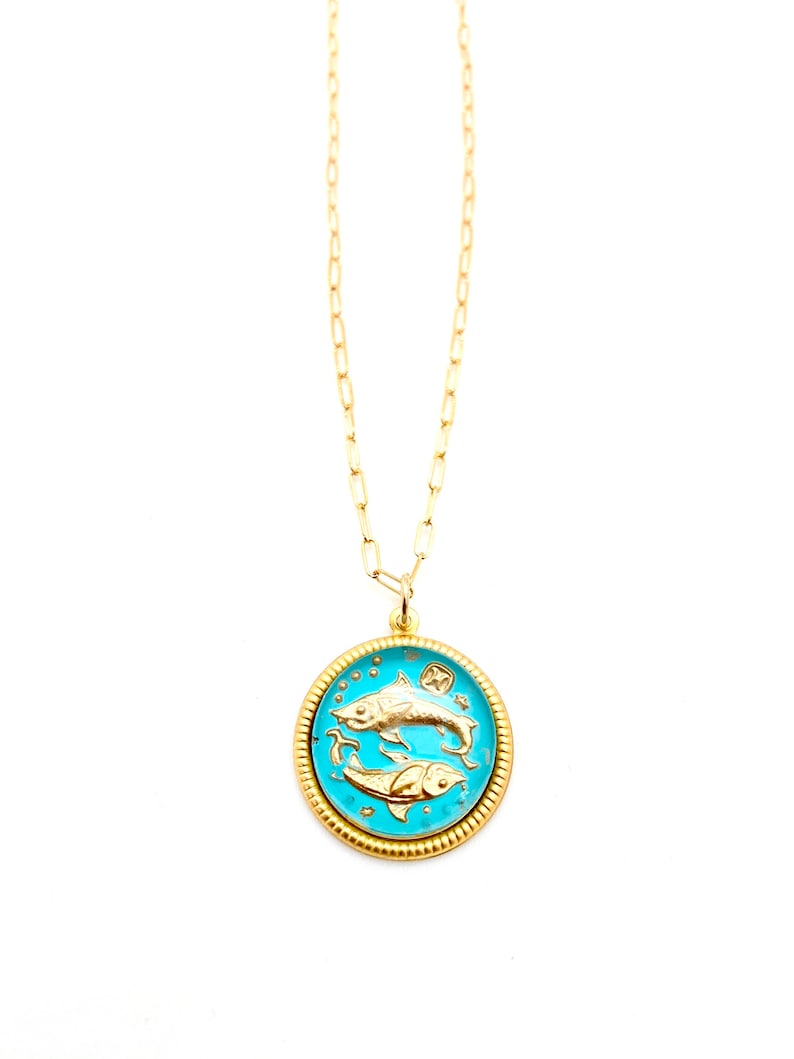 May include: A gold-tone necklace featuring a round pendant with a bright turquoise enamel inlay. The pendant displays two embossed gold fish swimming in opposite directions, representing the Pisces zodiac sign, with a textured gold border.