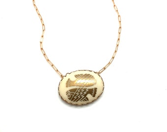 May include: A gold-toned chain necklace with an oval pendant. The pendant features a cream-colored background with two gold fish designs. The necklace is a delicate piece of jewelry.