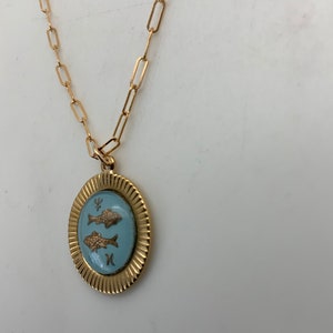 Puede incluir: Collar dorado con un colgante con un &oacute;valo azul claro con dos peces dorados. El colgante est&aacute; enmarcado por un borde dorado texturizado y cuelga de una cadena dorada con eslabones rectangulares. El collar se muestra sobre un fondo blanco.