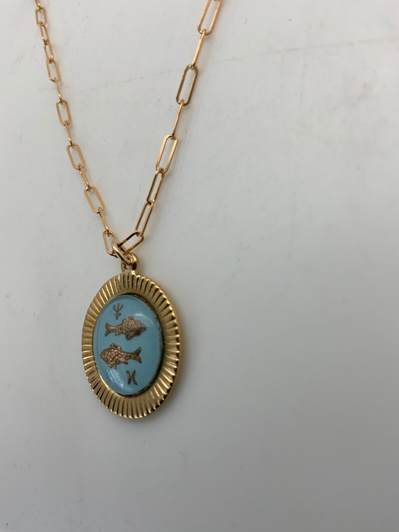 Puede incluir: Collar dorado con un colgante con un &oacute;valo azul claro con dos peces dorados. El colgante est&aacute; enmarcado por un borde dorado texturizado y cuelga de una cadena dorada con eslabones rectangulares. El collar se muestra sobre un fondo blanco.
