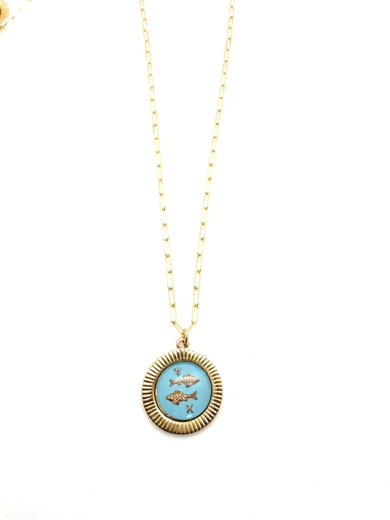 Puede incluir: Collar dorado con un colgante con dos peces sobre un fondo azul claro. El colgante es redondo con un borde texturizado. La cadena es delicada y cuelga sobre un fondo blanco.