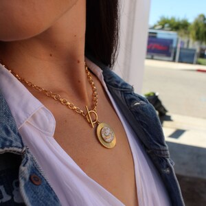 May include: A gold chain necklace with a large, round pendant featuring a white and gold design. The necklace is worn by a person with long dark hair and a white shirt.