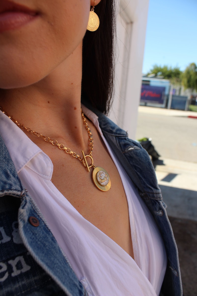 May include: A gold chain necklace with a large, round pendant featuring a white and gold design. The necklace is worn by a person with long dark hair and a white shirt.