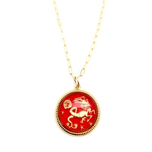 May include: A gold chain necklace featuring a round pendant with a red background. The pendant displays a gold-coloured embossed Capricorn zodiac symbol and small star accents, enclosed within a textured gold-toned circular frame.