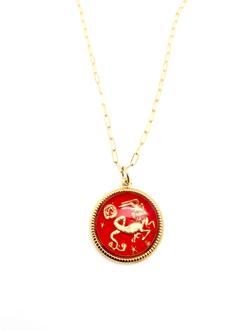May include: A gold chain necklace featuring a round pendant with a red background. The pendant displays a gold-coloured embossed Capricorn zodiac symbol and small star accents, enclosed within a textured gold-toned circular frame.