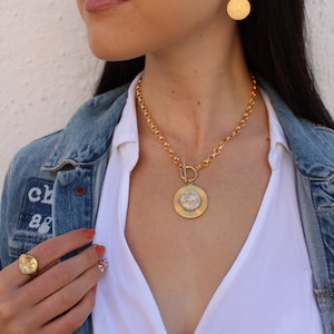 May include: A gold chain necklace with a large, round pendant featuring a textured design. The pendant is attached to the chain with a toggle clasp. The necklace is worn by a person with a white shirt and a denim jacket.