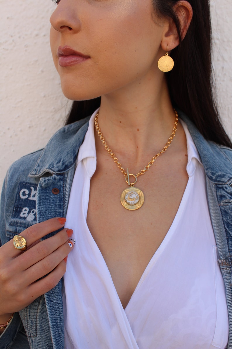May include: A gold chain necklace with a large, round pendant featuring a textured design. The pendant is attached to the chain with a toggle clasp. The necklace is worn by a person with a white shirt and a denim jacket.