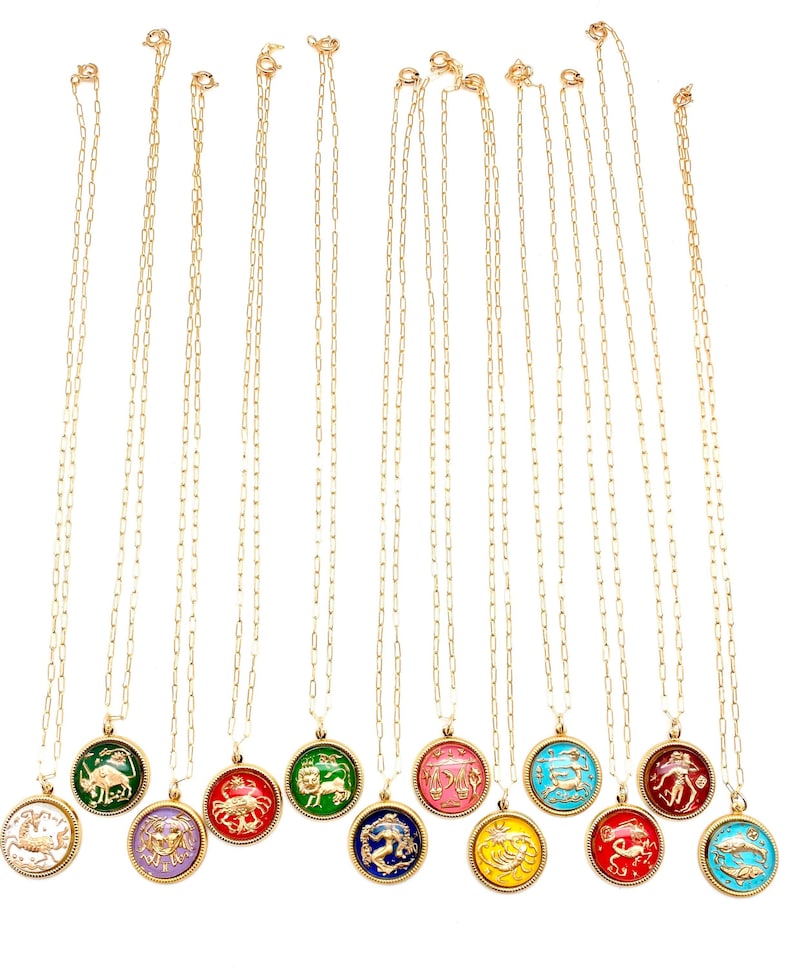 May include: A collection of twelve gold-tone chain necklaces, each featuring a round pendant with a raised zodiac symbol in gold against a vibrant, colourful enamel background. The pendants are suspended from delicate, elongated link chains.