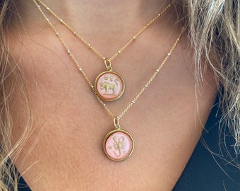 May include: Two gold necklaces with delicate chains and pink zodiac pendants. The top pendant features a bull, while the bottom pendant features a scorpion.