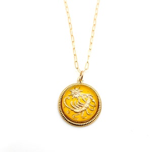 May include: A gold-toned necklace featuring a round pendant with a yellow enamel inlay. The pendant displays a raised gold scorpion figure and a small star. The piece hangs from a delicate gold-toned chain with elongated oval links.