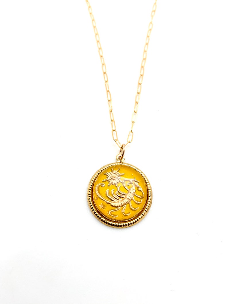 May include: A gold-toned necklace featuring a round pendant with a yellow enamel inlay. The pendant displays a raised gold scorpion figure and a small star. The piece hangs from a delicate gold-toned chain with elongated oval links.