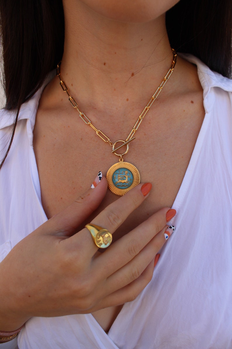 May include: A gold chain necklace with a large, round pendant featuring a blue enamel design. The pendant has a gold border and a raised design. The necklace has a toggle clasp.