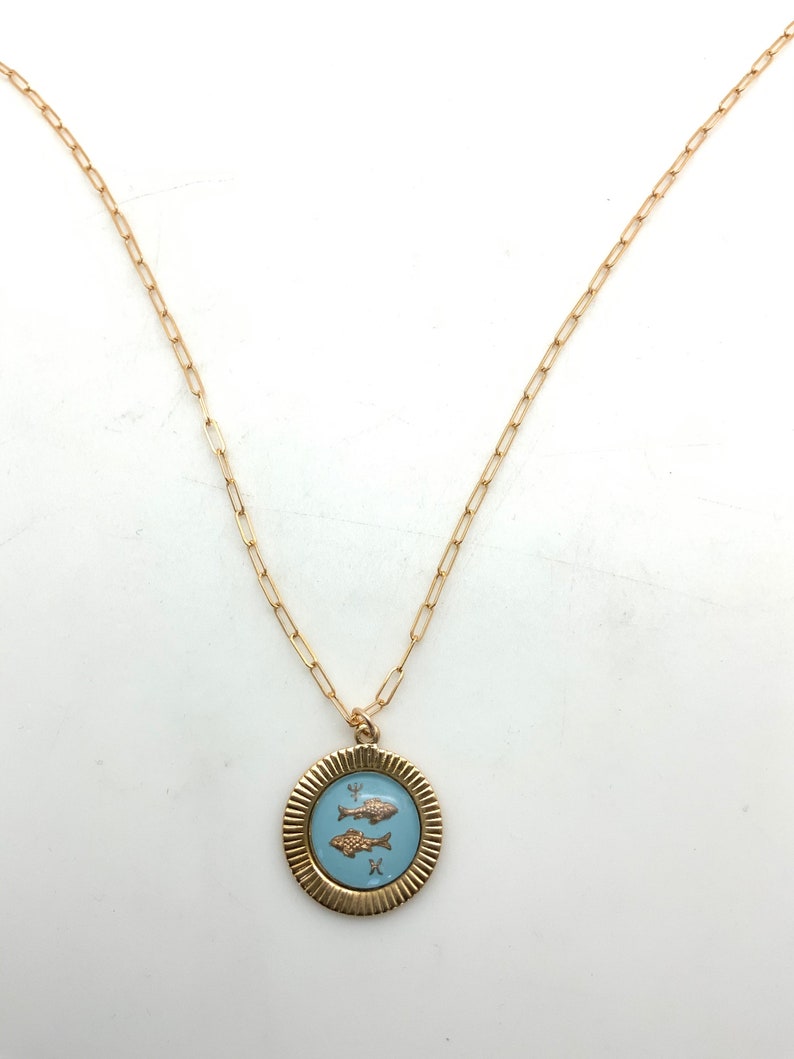 Puede incluir: Collar dorado con un colgante redondo con dos peces sobre un fondo azul claro. El colgante tiene un borde texturizado y acanalado. La cadena es de estilo eslab&oacute;n delicado.