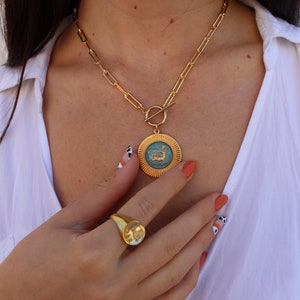 May include: A gold chain necklace with a large, round pendant featuring a blue enamel design. The pendant has a gold border and a raised design. The necklace has a toggle clasp.