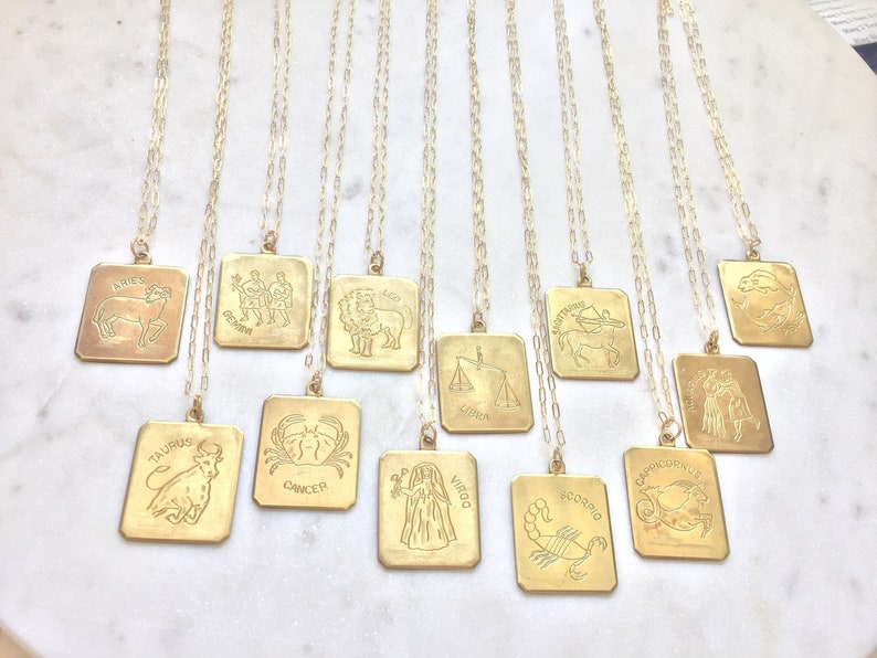 May include: Twelve gold necklaces with square pendants featuring zodiac signs. The signs include Aries, Taurus, Gemini, Cancer, Leo, Virgo, Libra, Scorpio, Sagittarius, Capricorn, Aquarius, and Pisces.