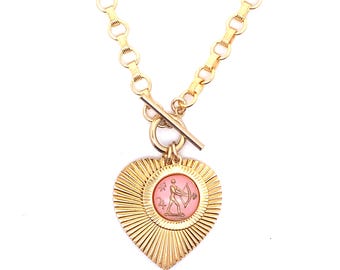 Op de afbeelding: Goudkleurige ketting met een hartvormige hanger en een sunburst design. De hanger heeft een roze centrum met een figuur die een pijl en boog vasthoudt. De ketting heeft een tuimelsluiting en een dikke ketting.