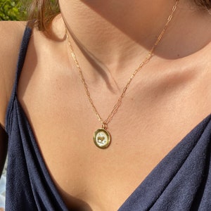 Puede incluir: Collar dorado con un colgante con un s&iacute;mbolo del zodiaco. El colgante es redondo con un fondo blanco y una ilustraci&oacute;n detallada. La cadena es delicada y cuelga del cuello de una persona, contra una parte superior azul oscuro.