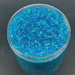 May include: A clear plastic container filled with small, rectangular, blue and teal beads. The beads have a reflective, iridescent quality, creating a shimmering effect. The container is set against a dark background.