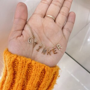 May include: A gold-toned necklace featuring a delicate chain with individual letter charms spelling the word TINKER. A small crescent moon charm encrusted with clear stones and a tiny fairy silhouette is attached to the left side of the chain.
