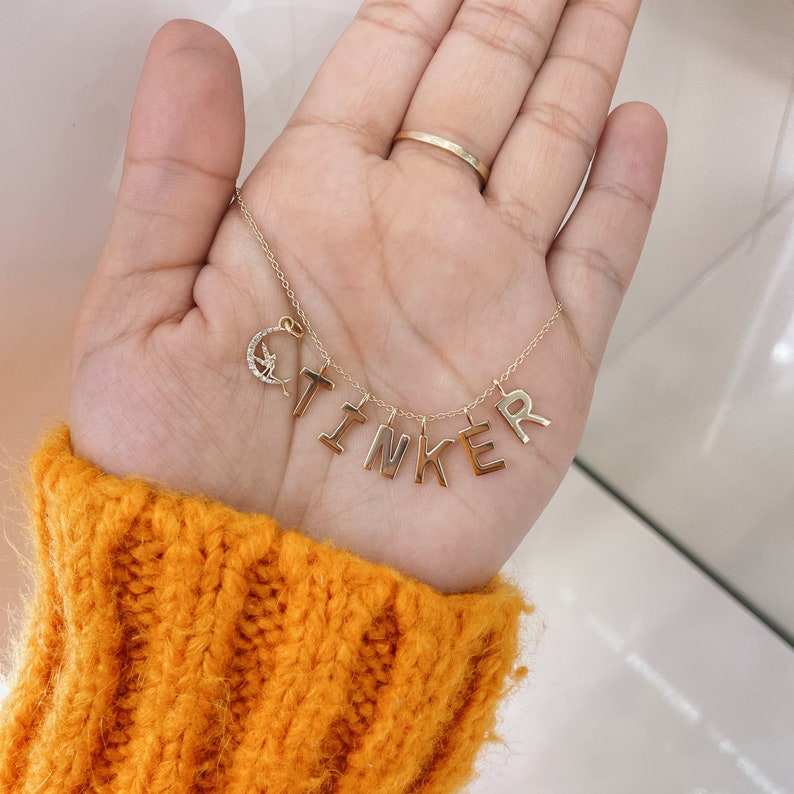 May include: A gold-toned necklace featuring a delicate chain with individual letter charms spelling the word TINKER. A small crescent moon charm encrusted with clear stones and a tiny fairy silhouette is attached to the left side of the chain.