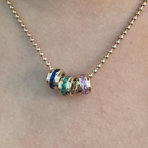May include: A gold chain necklace with four colorful rings. Each ring is set with small, square-shaped gemstones. The rings are blue, green, yellow, and pink.